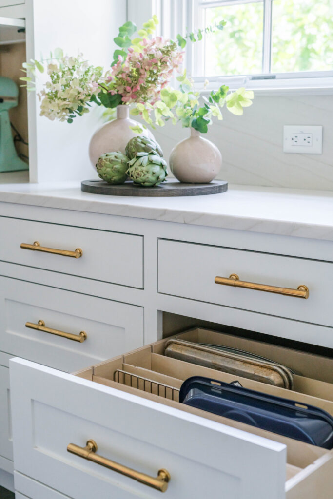 White kitchen with gold drawer handles, marble countertop, decorative vases, and open drawer with baking sheets.