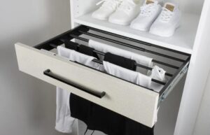 Open drawer with black metal rails holding folded clothes, beneath a shelf with two pairs of white sneakers.