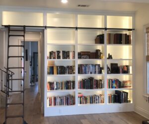 Large built-in bookshelf with assorted books, decorative items, sliding ladder, and soft backlighting in a modern room.