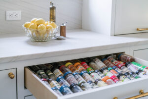 An open kitchen drawer displays organized spice jars; lemons in a glass bowl sit on the marble countertop above.
