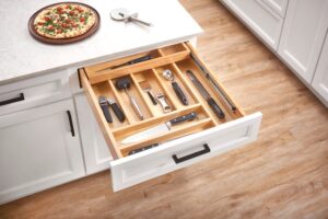 Open kitchen drawer with various utensils organized in wooden dividers; pizza and slicer on countertop above.